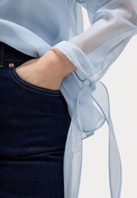 Blusa traslucida blu chiaro con maniche lunghe, legata in vita, abbinata a jeans in denim scuro con tasca frontale e dettaglio del bottone in ottone.