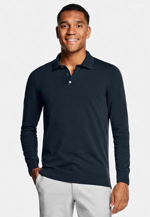 Lächelnder Mann, der ein dunkelblaues Langarm-Poloshirt und hellbeige Hosen trägt, mit einer Hand in der Tasche vor weißem Hintergrund stehend.