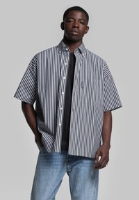 Camicetta a maniche corte con bottoni, a strisce verticali blu scuro e bianche, taschino sul petto, in tessuto di cotone, abbinata a una maglietta scura e jeans azzurri chiari.