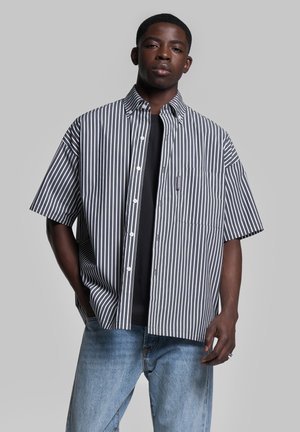 Button-up short-sleeve shirt with navy and white vertical stripes, chest pocket, cotton fabric, paired with dark t-shirt and light blue jeans.