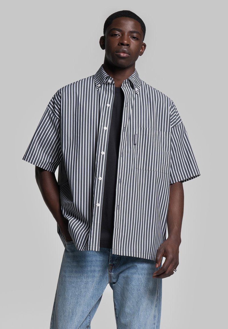 Camicetta a maniche corte con bottoni, a strisce verticali blu scuro e bianche, taschino sul petto, in tessuto di cotone, abbinata a una maglietta scura e jeans azzurri chiari.