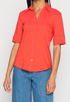 Nainen, jolla on kirkkaan punainen, lyhythihainen napillinen paita ja siniset korkeavyötäröiset farkut yksiväristä taustaa vasten.