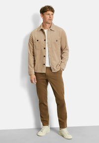 Beige Corduroy-Hemdjacke mit zwei Brusttaschen, kombiniert mit braunen Hosen und hellen Sneakers. Schlichtes Design, vertikale Textur.