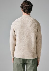 Pulover bej cu torsade, cu manșete raglan, având un model texturat de tricot. Asortat cu pantaloni verzi, văzut din spate.