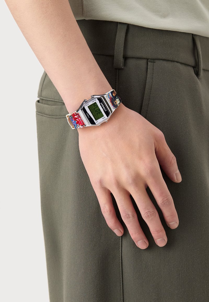 Reloj digital con una caja rectangular de color plateado, pantalla digital verde y una correa de diseño multicolor. Lleva puesto en una mano que descansa sobre un pantalón.