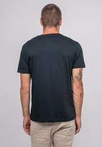 T-shirt noir à manches courtes en coton, avec un col rond et une texture lisse. Tatouage visible sur le bras gauche.