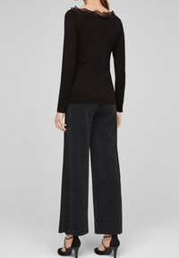 Kvinna iklädd en svart långärmad topp med volangkrage och glittriga svarta vida byxor, kompletterat med svarta högklackade skor.