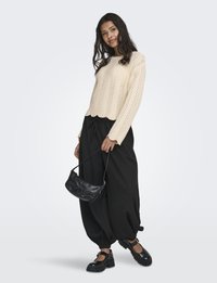 Jeune femme aux longs cheveux noirs portant un pull en tricot crème et un pantalon large noir, tenant un sac à main noir et portant des chaussures noires.