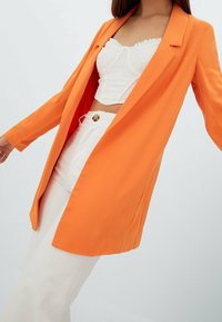 Blazer arancione con un design elegante e sartoriale, realizzato in tessuto liscio; indossato sopra una maglietta bianca aderente, abbinato a pantaloni bianchi.
