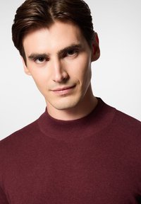 Burgundowa sweter z dzianiny z wysokim kołnierzem i prążkowanymi detalami, noszony przez mężczyznę, podkreślający dopasowany krój i gładką teksturę.