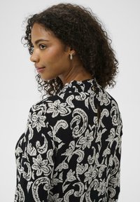 Must tekstuuriga top, millel on valged lillese ja rullmustri detailid, kõrge kaelusega ja tagaküljel võtmeauguga detail.