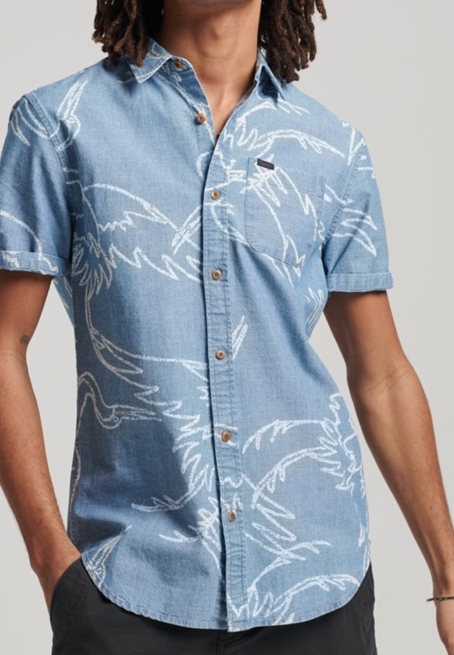 Homme portant une chemise bleu clair à manches courtes avec des motifs abstraits d'oiseaux blancs et un pantalon foncé, debout les mains dans les poches.