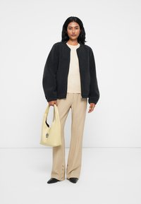 Veste zippée en fausse fourrure noire, haut beige, pantalon large beige clair avec boutons sur le côté, et un sac à main jaune doux. Chaussures noires à talons pointus.