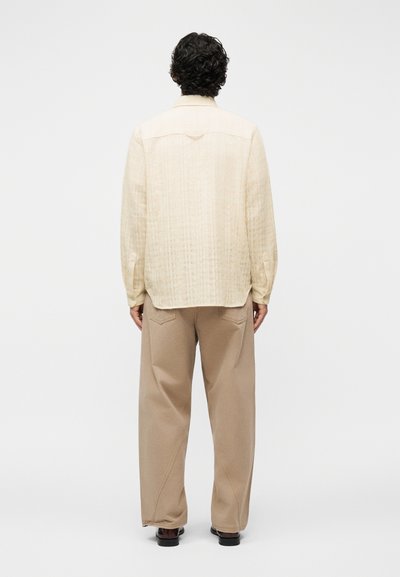 Άνδρας με σγουρά μαλλιά φορώντας μακρυμάνικο μπεζ πουκάμισο με υφή και άνετο ταμπά παντελόνι, στέκεται με την πλάτη προς τα εμπρός σε απλό φόντο.