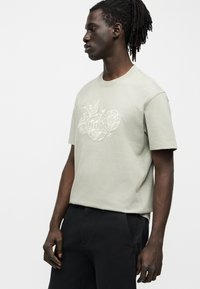 Homme avec dreadlocks portant un t-shirt gris clair avec une broderie florale et le logo "HUGO", associé à un pantalon noir, debout de profil.