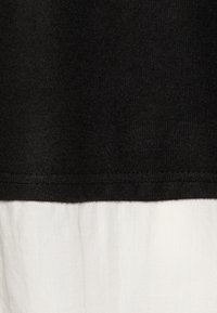 Svart tygtop med en texturerad yta och rak skärning, lager på en slät vit underdel. Synlig sömnad längs fållen.