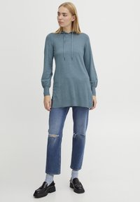 Tunik s dolgim rokavima in kapuco v teal barvi, izdelana iz mehkega materiala. Ima rebraste manšete in sproščen kroj. Oblečena je z unikatnimi modrimi kavbojkami in črnimi mokasini.