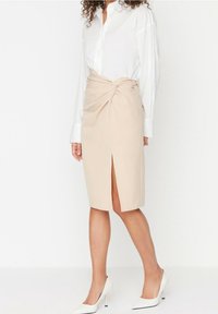 Jupe crayon beige avec une fente sur le côté et un détail noué à la taille, associée à une chemise blanche à boutons et des chaussures à talons blancs.