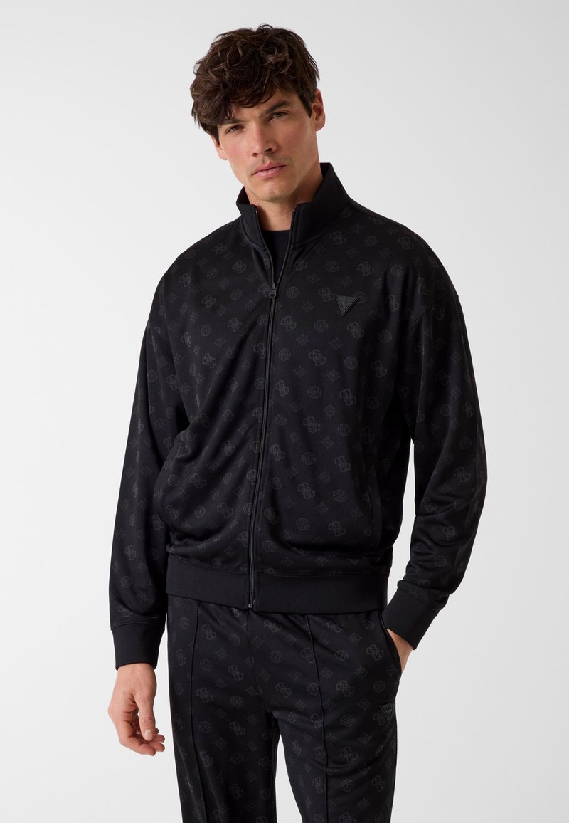 Mann mit dunklem Haar, der eine schwarze Zip-up-Jacke und passende Hose mit dezentem, wiederholtem Logo-Muster trägt, posiert vor weißem Hintergrund.