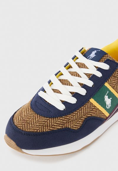 Baskets bleu marine et marron avec un tissu chevron, lacets blancs, accent vert et jaune, et un patch logo. Semelle en caoutchouc avec un détail texturé.