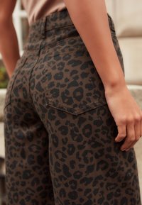 Pantalons en imprimé léopard marron et noir. Design taille haute avec une texture lisse et des coutures décoratives sur les poches. Main reposant sur la hanche.