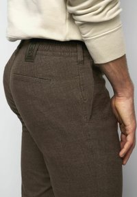 Nahaufnahme einer Person, die braune, strukturierte Hosen mit einer Seitentasche und ein beiges Langarmoberteil trägt, wobei ein Teil der rechten Hand zu sehen ist.