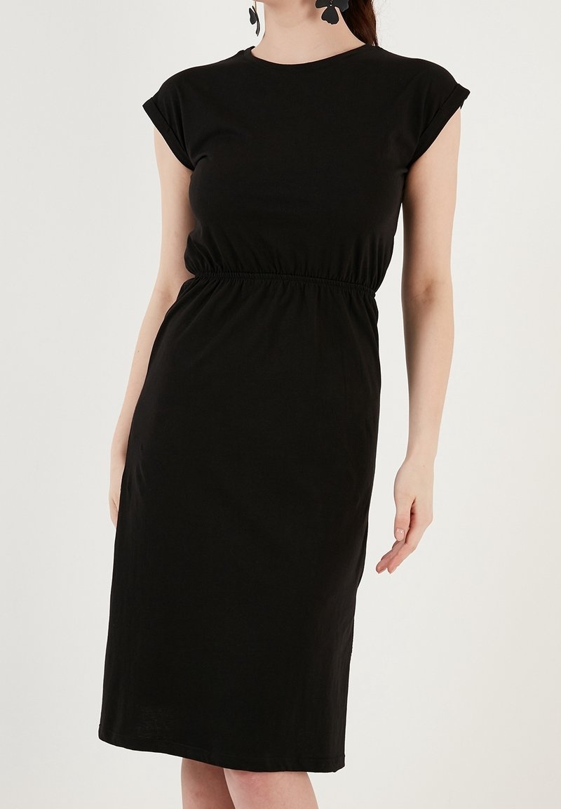 Femme portant une robe noire sans manches d'une longueur au genou avec une taille plissée et des boucles d'oreilles florales noires sur un fond clair uni.