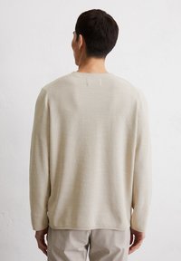 Beige gebreide trui met lange mouwen, textuurstof, ronde hals en geribde manchetten, gedragen over lichtgekleurde broeken.