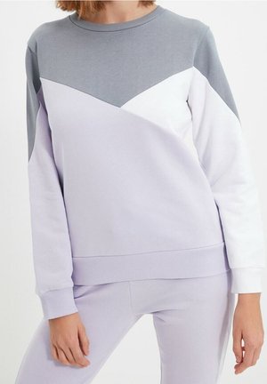 Femme portant un sweat-shirt bicolore avec des sections grises, blanches et violet clair, assorti à un pantalon violet clair.