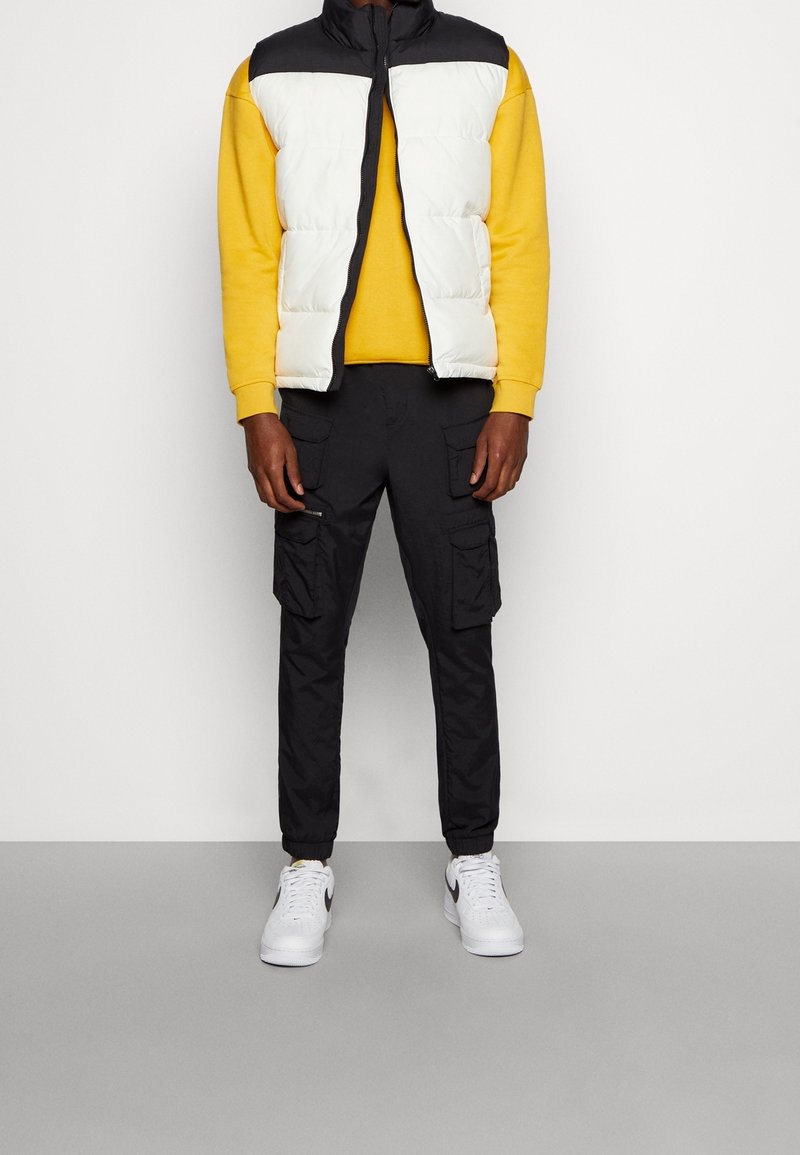 Homme portant un sweat-shirt jaune, une veste matelassée blanche et noire, un pantalon cargo noir et des baskets blanches, debout devant un fond uni.