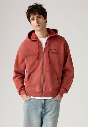 Rode hoodie met rits, een trekkoordkap, een oversized ontwerp en voorzakken. Heeft "TWO HORSE ATHLETIC CLUB" in donkerbruine letters.