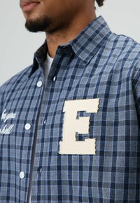 Blauw geruit overhemd van geweven stof. Heeft een beige geborduurde letter 'E' op de borst en witte tekst aan de linkerkant.