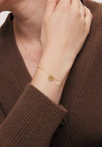 Złota bransoletka zdobiona dwoma charmsami w kształcie kwiatów, jednym szczegółowym i jednym minimalistycznym, noszona na nadgarstku, uzupełniona brązowym swetrem z dzianiny.