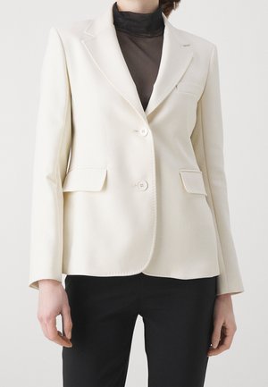 Vrouw draagt een crèmekleurige dubbelgeknoopte blazer over een zwart doorschijnend hooggesloten topje en zwarte broek, staand tegen een effen achtergrond.