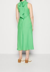 Zielona sukienka bez rękawów z tied kokardą przy szyi, wykonana z gładkiego materiału o długości midi. Noszona z białymi klapkami na szerokim obcasie.