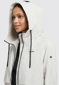 Lichtgrijze hooded jas met zwarte trekkoorden, ritssluiting en subtiele logo-details. Gladde textuur, middellange ontwerp en functionele capuchon.