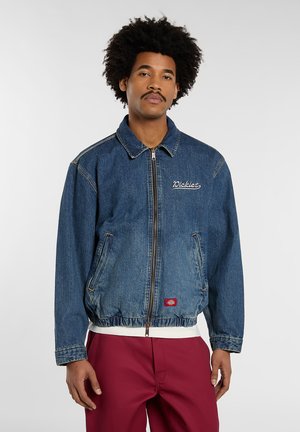 Homme aux cheveux bouclés portant une veste en jean bleue zippée et un pantalon rouge, debout sur un fond uni.