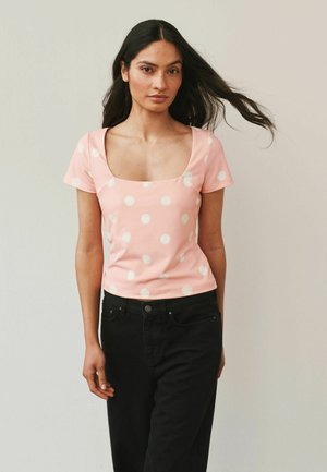 Hosszú sötét hajú nő rózsaszín, fehér pöttyös rövid ujjú felsőt és fekete nadrágot visel, egyszínű háttér előtt áll.