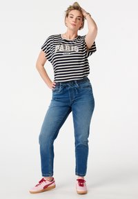 T-shirt rayé noir et blanc avec le texte "PARIS", associé à un jean bleu à taille élastique et des baskets roses. Le modèle pose avec une main sur la hanche.