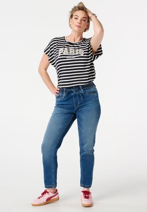 Gestreepte zwart-witte T-shirt met de tekst "PARIS", gecombineerd met blauwe jeans met elastische taille en roze sneakers. Model poseert met één hand op de heup.