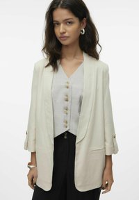 Kreem värvi linane bleiser, millel on šaalikaelus, kaks eesmist taskut ja rullitud varrukad, kantud halli nööpidega pluusi ja mustade pükste üle.