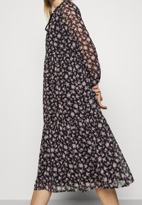 Robe noire à fleurs avec des manches longues transparentes, un corsage ajusté et une jupe évasée, ornée de petits motifs floraux roses et blancs.