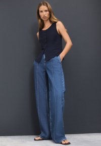 Kvinde står med hænderne i lommerne iført ærmeløs navyblå knappet top, vidbenede blå jeans med syninger foran og sorte sandaler foran en grå væg.