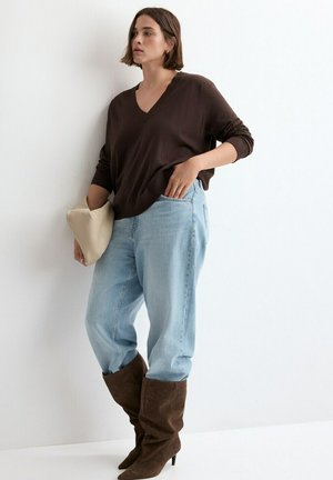 Femme portant un pull ample marron à col en V, un jean bleu clair ample et des bottes marron souples montant jusqu'aux genoux, tenant une pochette beige sur un fond blanc.