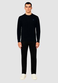 Maglione blu a maglia con collo rotondo, abbinato a pantaloni neri e sneaker bianche. Mostra un logo blu sul petto.