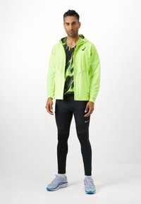 Giacca leggera verde neon con cappuccio, top senza maniche nero a motivi, leggings neri e scarpe da ginnastica azzurro chiaro con accenti grigi e logo.
