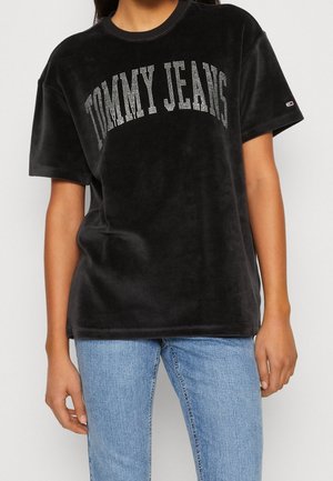 Henkilö, joka pukee mustan veluuripaidan, jossa lukee "TOMMY JEANS", ja vaaleansiniset farkut, seisoo yksiväristä taustaa vasten.