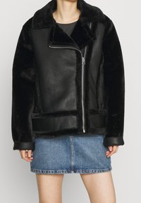 Kvinna iklädd en svart jacka i konstläder och fårskinn med asymmetrisk dragkedja, kombinerad med en blå denim minikjol.