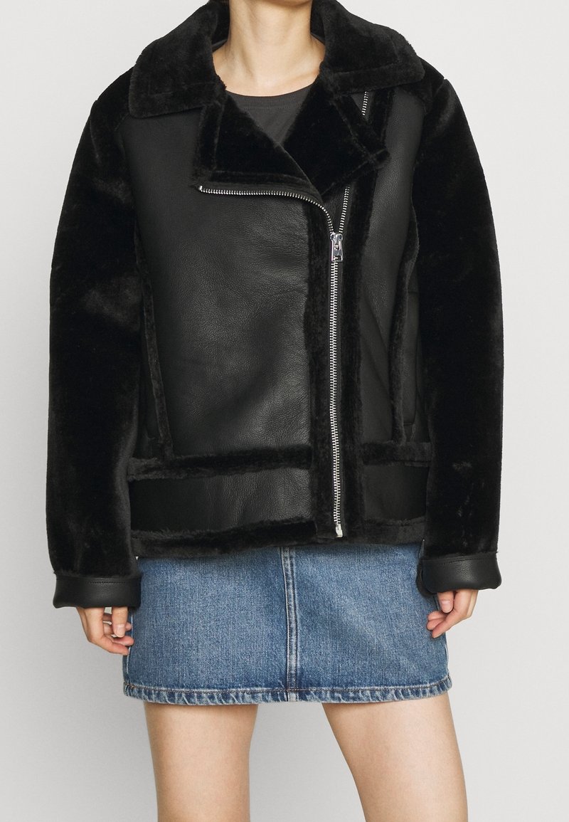 Kvinna iklädd en svart jacka i konstläder och fårskinn med asymmetrisk dragkedja, kombinerad med en blå denim minikjol.