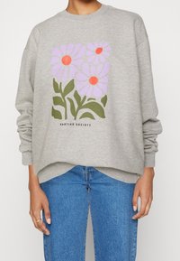 Persona che indossa un felpa grigia con tre fiori viola con il centro rosso e gambi verdi, abbinata a jeans blu.